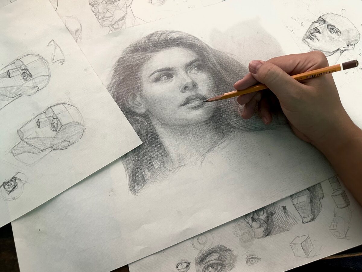 A hand works on a shaded portrait drawing on paper